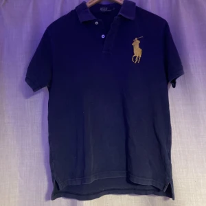Mörkblå Polo Ralph Lauren t-shirt - Snygg mörkblå t-shirt från Polo Ralph Lauren med klassisk polokrage och två knappar. Stor gul broderad logga på bröstet och gul detalj på ärmen. Tillverkad i mjuk bomull, perfekt för en chill och stilren look.