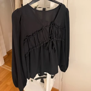 Svart blus med volang och knyt - Säljer en svart blus med lång ärm och v-ringning. Blusen har en bred volangdetalj tvärs över bröstet och knyt framtill. Materialet är lätt. Aldrig använd.