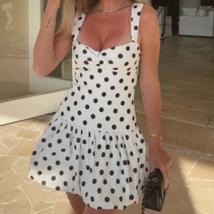 Vit prickig klänning från Zara - Supersöt vit klänning från Zara med svarta prickar, breda axelband och en lätt puffig ballong kjol. Klänningen har en snygg v-ringad överdel med detalj och dragkedja i ryggen. 