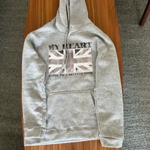 Grå hoodie med brittisk flagga - Säljer en grå hoodie med tryck av brittiska flaggan och texten 'MY HEART BEATS TO A BRITISH TUNE' på bröstet. Tröjan har huva med snörning, magficka och är tillverkad i mjuk polyester. Perfekt för dig som gillar streetstyle och vill sticka ut.