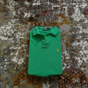 Grön pikétröja från Polo Ralph Lauren - Snygg grön pikétröja från Polo Ralph Lauren i slim fit-modell. Klassisk krage, kort ärm och gul broderad logga på bröstet. Tillverkad i bomull som känns skön mot huden. Perfekt för dig som gillar stilrena och sportiga vibes.