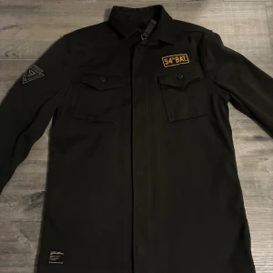 Mörkgrön overshirt från Superdry S - Mörkgrön overshirt från Superdry med coola patchar på bröstet och ärmen. Jackan har två bröstfickor med knappar och klassisk krage. Perfekt för lager-på-lager och streetstyle. Modellen är rak och normal i passformen.