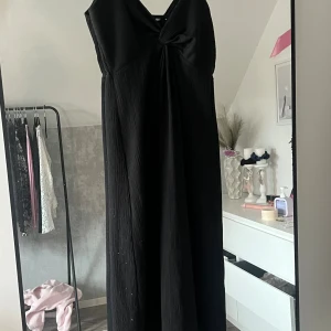 Svart långklänning med twistad front - Elegant svart långklänning med v-ringning och twistad detalj framtill. Klänningen har en mjukt fallande siluett och är ärmlös, perfekt för en stilren look. Materialet ser ut att vara lätt och följsamt, vilket ger en snygg drapering.