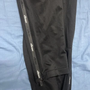 Svarta mjukisbyxor från Ellesse - Svarta mjukisbyxor från Ellesse med elastisk midja och dragkedjor längs benen med logotyp. Snygg sportig look och bekvämt material som passar perfekt för chill eller träning. Klassisk design med raka ben och diskreta fickor.