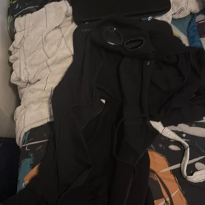 Svarta kofta från CP Company - Svart kofta från CP Company med dragkedja och snörning i midjan. Kofta har en clean look och är gjorda i ett slitstarkt syntetmaterial, perfekta för kalla dagar. Snyggt och funktionellt plagg för dig som gillar streetwear och vill ha något som sticker ut.