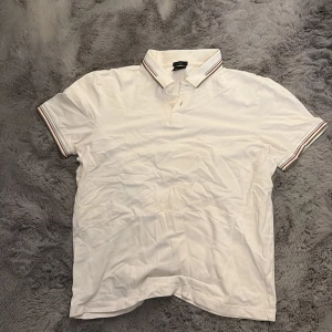 Vit slim fit piké från Hugo Boss - Vit piké från Hugo Boss i slim fit med klassisk krage och korta ärmar. Kragen och ärmsluten har snygga ränder i rött och beige som ger en sportig vibe. Tillverkad i mjuk bomull för skön känsla hela dagen.