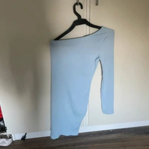 Ljusblå one shoulder klänning - Snygg ljusblå klänning med one shoulder-design och lång ärm. Klänningen är ribbad och har en kort längd som ger en trendig och ungdomlig vibe. Perfekt för dig som gillar stilrena och moderna plagg med en twist. Använd en gång på en skoldans.