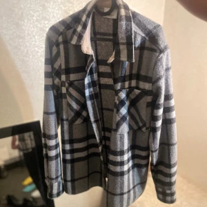 Rutig overshirt från Burberry - Snygg rutig overshirt från Burberry i grått, svart och vitt. Skjortan har klassisk krage, två bröstfickor med lock och knappar framtill. Materialet är mjukt och värmande ull, perfekt för lager-på-lager. Den har långärmad passform och stilren design. Storlek s men passar m 