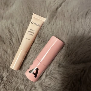 CAIA That Dewy Look & Cocoa Couture Lip Balm - Säljer två sminkprodukter från CAIA: en beige tub med 'That Dewy Look Eye Gloss' för glansiga ögonlock och en ljusrosa hylsa med 'Cocoa Couture Lip Balm' som ger mjuka läppar. Stock endast kikat färg 