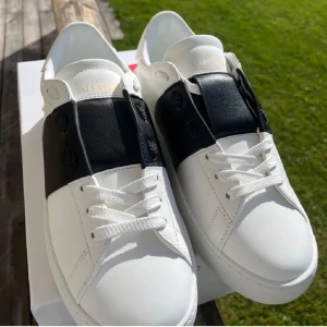 Valentino Garavani Open sneakers vit/svart - Snygga Valentino Garavani Open sneakers i vitt skinn med bred svart läderdetalj över snörningen. Platt sula med mönstrad undersida och ikoniska nitar baktill. Klassisk rund tå och vita skosnören. Perfekt för dig som gillar stilrena designer med lyxig känsla.