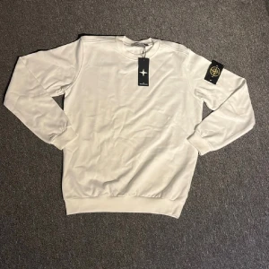 Vit sweatshirt från Stone Island - Vit sweatshirt från Stone Island med klassisk rund hals och avtagbar logotyp-patch på ärmen. Tröjan har en clean look med ribbade muddar och är tillverkad i mjuk bomull. Perfekt för dig som gillar stilrena och trendiga plagg.