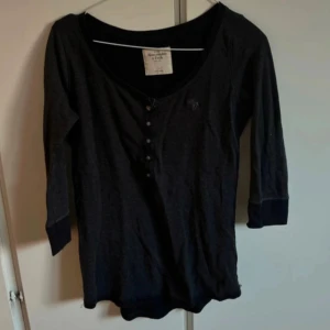Svart topp från Abercrombie & Fitch - Svart topp från Abercrombie & Fitch med trekvartsärm och rund halsringning. Framsidan har en knappslå med små knappar och en diskret broderad logga på bröstet. Perfekt för en chill och stilren look.