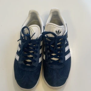 Adidas Gazelle sneakers marinblå/vita - Säljer ett par klassiska Adidas Gazelle sneakers i marinblå mocka med vita detaljer och de ikoniska tre ränderna på sidorna. Skorna har snörning, vit sula och Adidas-logga på häl och plös. Perfekta för dig som gillar retrostil och streetwear.