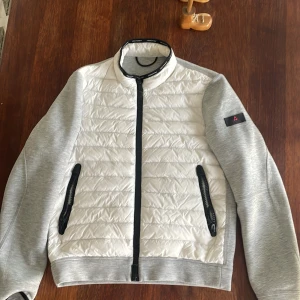 Vit och grå hybridjacka från peuterey - Snygg hybridjacka från peuterey  med quiltad vit front och grå ärmar och rygg. Jackan har svarta dragkedjor, ståkrage och två fickor framtill. Perfekt för dig som gillar sportig och stilren look peuterey-logga på ärmen. Original pris-5000kr
