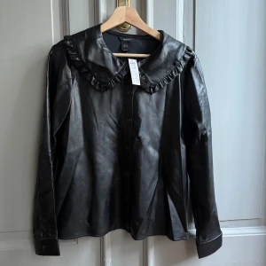 Svart skjorta i skinnimitation Lindex M - Svart skjorta från Lindex i skinnimitation med stor krage och volangdetalj. Långa ärmar, knäppning framtill. 