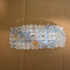 Blommig hårklämma med pärlor - Superfin hårklämma dekorerad med små blommor i ljusblå, vit och beige nyans, varje blomma har en glittrande pärla i mitten. Klämman har en rektangulär form och metallspänne på baksidan. Perfekt för att ge frisyren en söt och unik touch.