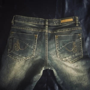 Blå jeans från Lab Industries - Säljer ett par blå jeans från Lab Industries med slitna detaljer och kontrastsömmar. Bakfickorna har broderade mönster i gult och nitar. 