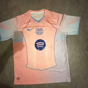 Barcelona fotbollströja Nike rosa/blå - Säljer en FC Barcelona fotbollströja från Nike i en snygg ljusrosa och ljusblå färgkombination. Tröjan har klubbmärke, Nike-logga och Spotify som sponsortryck på bröstet. Tillverkad i lätt och ventilerande material, perfekt för match eller träning.