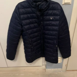 Mörkblå pufferjacka från Gant - Snygg mörkblå pufferjacka från Gant med horisontella quiltade paneler och hög krage. Jackan har dragkedja framtill och två fickor med knappstängning. Perfekt för kyliga dagar och har en stilren look som funkar till det mesta. Pris kan diskuteras vid snabb affär.