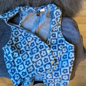 Blommig jeanströja från H&M Divided - Säljer en blå jeanströja från H&M Divided med blommigt mönster i olika blå nyanser. Västen har krage, tryckknappar framtill och är figursydd. Kan ha något under men går lika bra utan.