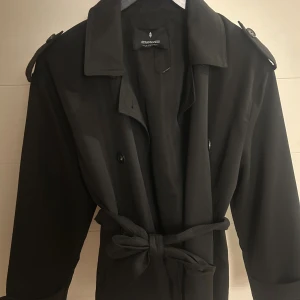Svart trenchcoat från Stradivarius - Svart trenchcoat från Stradivarius med klassisk krage, knappar framtill och bälte i midjan. Jackan har axelklaffar och detaljerade ärmslut med knappar. Perfekt för dig som gillar stilrena och tidlösa plagg.