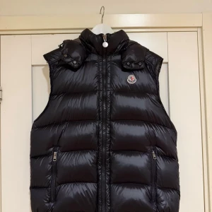 Svart dunväst från Moncler - Säljer en svart dunväst från Moncler med glansig finish och hög krage. Västen har två praktiska fickor med dragkedja och ikonisk Moncler-logga på bröstet. Insidan har ett unikt serietidningsmönster och märkets autenticitetsmärkning. Perfekt för att lyfta din streetstyle. Storlek 3= M 