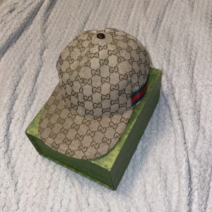 Beige keps från Gucci - Säljer en snygg beige keps från Gucci med det klassiska GG-mönstret. Kepsen har en justerbar rem i skinn och en grön-röd rand på sidan. Perfekt för att ge din outfit en lyxig touch. Kepsen är i mycket bra skick och kommer med originalförpackning.