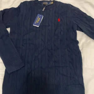 Marinblå stickad tröja Polo Ralph Lauren - Snygg marinblå stickad tröja från Polo Ralph Lauren med slim fit-passning. Tröjan har kabelstickat mönster, rund hals och den klassiska röda Polo-loggan broderad på bröstet. Perfekt för dig som gillar stilrena och tidlösa plagg.