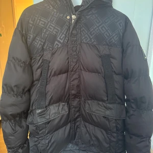 Svart pufferjacka Glorious Gangsta M - Svart pufferjacka från Glorious Gangsta med mönstrade paneler upptill och på huvan. Jackan har stora fickor framtill, dragkedja och tryckknappar, luvan har med sig pälsen också. 