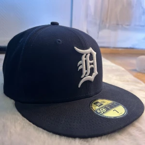 Svart New Era 59FIFTY Detroit Tigers keps - Snygg svart 59FIFTY keps från New Era med Detroit Tigers-logga i vitt framtill. Klassisk flat brim och broderade MLB- och New Era-loggor på sidan och bak. Officiell on-field cap, perfekt för dig som gillar sportig streetstyle.
