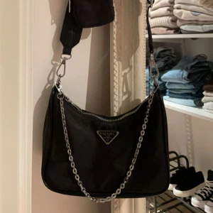 Svart axelväska från Prada med kedja - Snygg svart axelväska från Prada med silverfärgad kedja och extra liten ficka på axelremmen. Väskan har klassisk trekantig Prada-logga i metall framtill och dragkedja upptill. Materialet är slitstark nylon och remmen är justerbar för perfekt passform. Jättefint skick! 