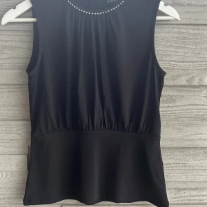 Svart pärldekorerad topp från Stockh LM - Snygg svart topp från Stockh LM i storlek XS. Modellen är ärmlös med rund halsringning dekorerad med vita pärlor. Toppen har en lätt rynkad midja och slät nederdel. Toppen är oanvänd.