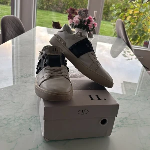Valentino Rockstud Untitled sneakers - Snygga Valentino Rockstud Untitled sneakers i vitt skinn med svarta detaljer och ikoniska nitar över vristen. Klassisk rund tå, platt sula och snörning framtill. Skosnörena är lite smutsiga men med ett par nya så ser mycket fräschare ut. Dom är använda men är fortfarande ett mycket  bra shick men har ett litet hål under skon men ser knappt det. 
