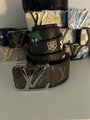 svart Läder bälte med monogram - Snyggt svart bälte med klassiskt monogrammönster och logga i silver på spännet. Bältet har detaljer i gult och blått med texten 'Vuitton' på sidan. Perfekt accessoar för att lyfta din outfit.