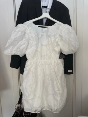 Vit kort klänning med puffärmar - Supersöt vit kort klänning med puffiga ärmar och blommigt mönster. Klänningen har öppen rygg med knyt och voluminös siluett. Lappen är kvar på och aldrig använd, bara testat 1gång☺️💕