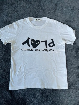 Vit Comme des Garçons PLAY t-shirt - Snygg vit t-shirt från Comme des Garçons PLAY med det ikoniska hjärtlogotyp-trycket och svart text på bröstet. 