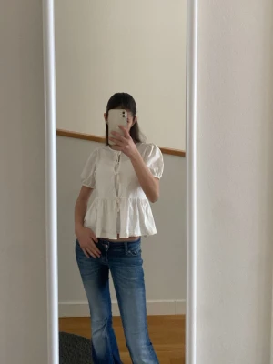 Vit blus med knyt och puffärm - Söt vit blus med korta puffärmar och knyt framtill. Blusen har en lös, volangig passform och rund halsringning. Perfekt för en trendig och avslappnad look. Materialet ser ut att vara bomull eller en bomullsblandning.