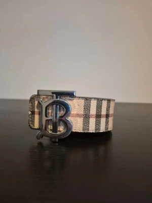 Beige och svart Burberry bälte - Snyggt bälte från Burberry med klassiskt beige och svart rutigt mönster och en stor metallspänne med Burberrys B-logga. Perfekt accessoar för att lyfta din outfit med en ikonisk touch.