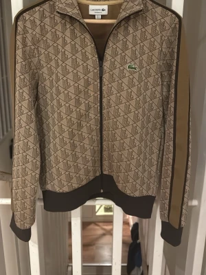  Lacoste ziptröja  - Säljer en beige ziptröja från Lacoste med geometriskt mönster och bruna detaljer. Tröjan har hel dragkedja, ståkrage och den klassiska gröna krokodilloggan på bröstet. Ribbade muddar och en brun rand längs ärmarna ger en sportig touch.
