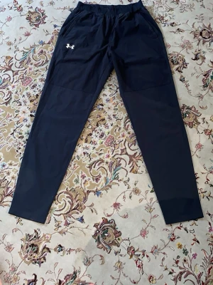 Svarta träningsbyxor från Under Armour - Säljer ett par svarta träningsbyxor från Under Armour med elastisk midja och två sidofickor. Byxorna har en vit logga på vänster lår och reflekterande detaljer vid bensluten. Perfekta för löpning eller gympass.