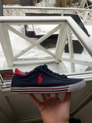 Marinblå Polo Ralph Lauren sneakers - Snygga marinblå sneakers från Polo Ralph Lauren med röd logga på sidan, röda detaljer vid hälen och röd sula. Skorna har vita och röda ränder runt sulan och klassisk låg profil. Perfekta för en stilren och sportig look.
