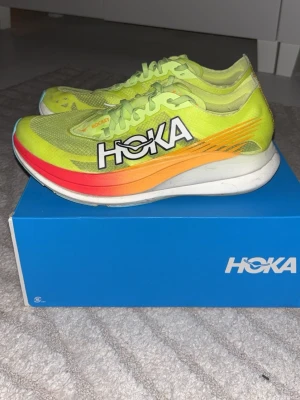Hoka Rocket X2 löparskor (kolfiber) - Säljer ett par Hoka Rocket X2 löparskor i en riktigt snygg neongul färg med orange och röd gradient längs sidan. Skorna har ventilerande mesh-överdel och tydlig HOKA-logga på sidan. Dojjan har även en kolfiberplatta vilket gör det till en perfekt tempo/tävlingssko, anpassad för maximal hastighet. Perfekta för dig som vill springa snabbt och sticka ut på löprundan. Storlek 42😁 köpta på löplabbet för 3300kr, kvitto finns!