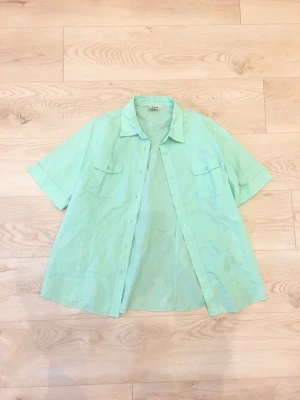 Ljusgrön kortärmad skjorta - Snygg ljusgrön kortärmad skjorta. Tyget är skönt luftigt bäckebölja. Skjortan har två bröstfickor med lock och knappar framtill. Perfekt som en layering piece för vår och sommar. 