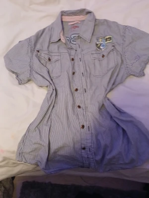 shirt for sale size L/Xl - Säljer en blåvit randig skjorta med korta ärmar, bröstfickor och broderade detaljer på framsidan. Skjortan har klassisk krage, knappar framtill och en avslappnad passform. Perfekt för dig som gillar sportig och casual stil.