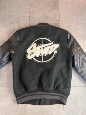 Svart bomberjacka från SWDZ - Svart bomberjacka med stora broderade SWDZ-loggor på ryggen och bröstet. Jackan har ärmar i svart skinnimitation och kropp i ullblandning, ribbade muddar och tryckknappar framtill. Snygga detaljer med brodyr och patchar på ärmen.