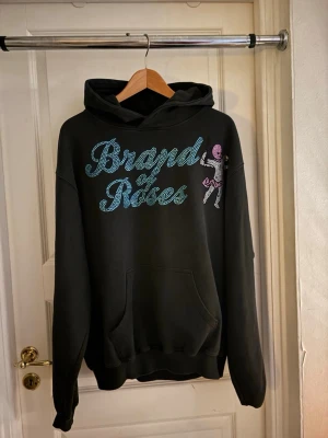 Svart hoodie Brand of Roses - Svart hoodie från Brand of Roses med stor ficka framtill och huva. Framsidan har ett stort tryck i blått och grönt med texten 'Brand of Roses' samt en liten figur i rosa och vitt. Perfekt för dig som gillar streetwear och coola prints.