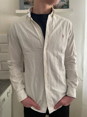 Vit skjorta från Ralph Lauren - Klassisk vit skjorta från Ralph Lauren med broderad logga på bröstet. Skjortan har lång ärm, knappar framtill och en smal passform. Tillverkad i bomull och passar perfekt till en stilren look.