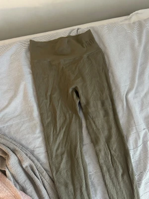 Olivgröna seamless tights från stronger  - Säljer ett par olivgröna seamless tights från stronger. De har hög midja, ribbade detaljer och bred linning för extra stöd. Materialet är stretchigt och följsamt, perfekt för träning eller gym. Snygga detaljer vid låret och diskret logga på linningen.
