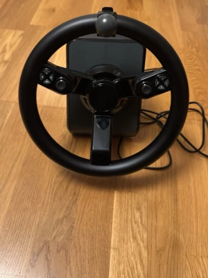 Logitech ratt och kontrollpanel - Säljer ett svart racingrattset och kontrollpanel från Logitech. Ratten har flera knappar och är i plast med en klassisk rund form. Kontrollpanelen har färgglada knappar i lila, orange, gul, blå och grön samt joystick och vred. Perfekt för simracing eller farming simulator.