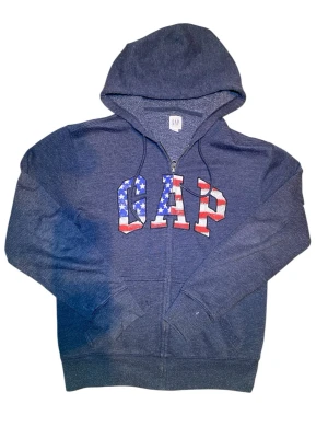 Mörkblå GAP hoodie med USA-flagga - Mörkblå zip-hoodie från GAP med stor logga på bröstet i USA-flaggsfärger. Hoodien har dragsko i huvan, två fickor fram och ribbade muddar. Tillverkad i mjukt bomullsmaterial, perfekt för en avslappnad streetwear-look.